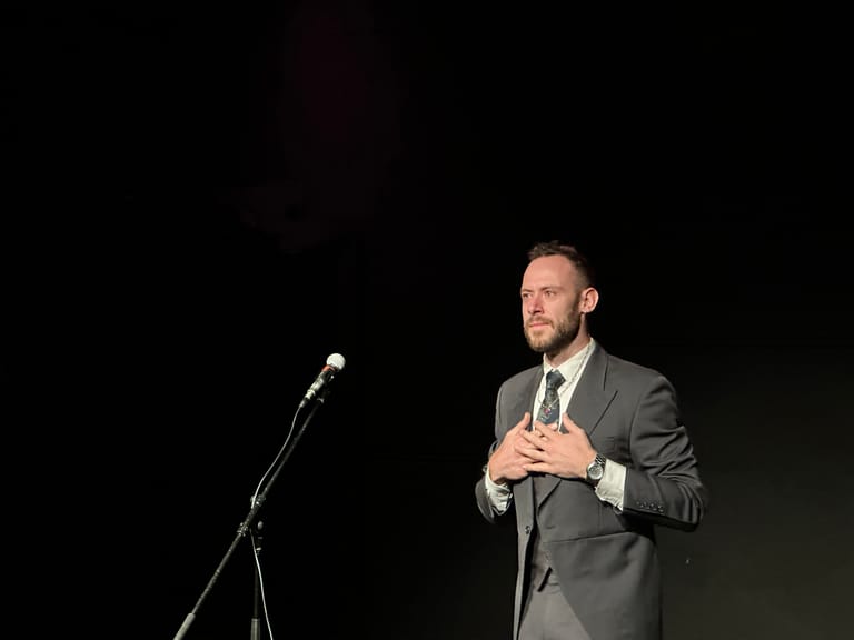 Man in grey suit speaking at microphone on darkened stage.