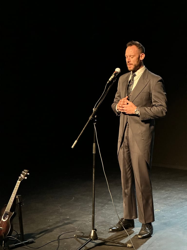 Comedian in a grey suit speaking at a microphone on a darkly lit stage with a guitar nearby.