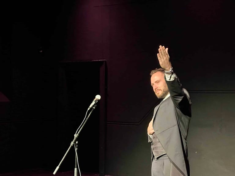 Performer in grey suit raises hand to audience beside microphone on darkened stage.