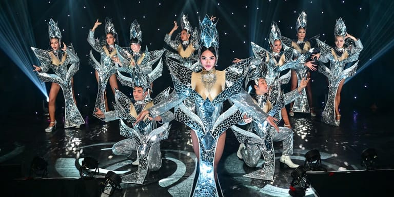 Performers in sequined costumes and headdresses strike poses on stage under dramatic blue lighting during The Ladyboys Of Bangkok show.