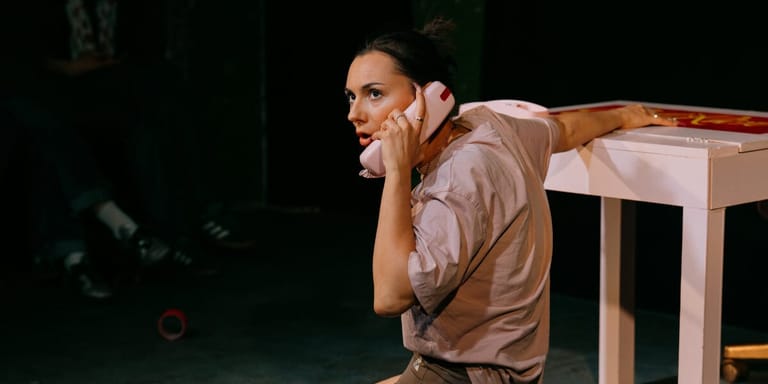 Performer in a brown shirt leans against a wooden table on stage, speaking expressively into a microphone.