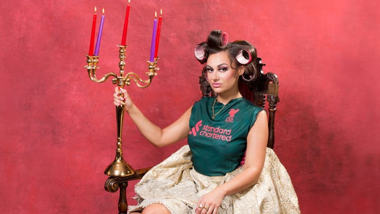 Performer in green top and cream skirt holds ornate golden candelabra against bright red backdrop.