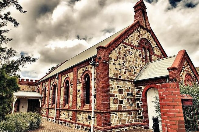 Historic brick and stone church building with arched windows and a tall chimney against a cloudy sky.
