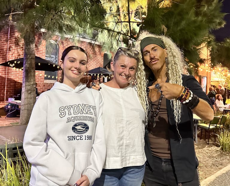 Three performers pose together outdoors at night in front of brick buildings and string lights.