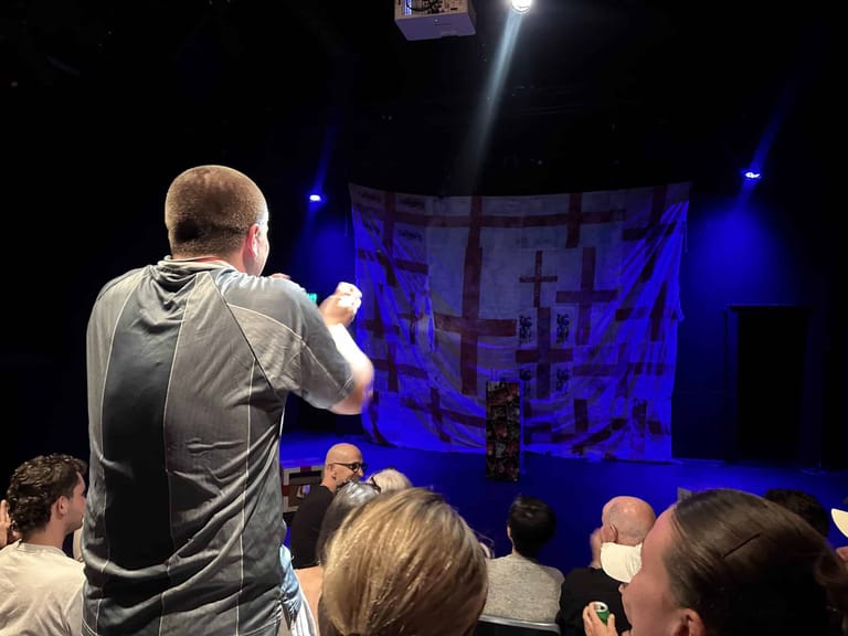 Performer on stage addressing an audience seated in the foreground, bathed in blue stage lighting.