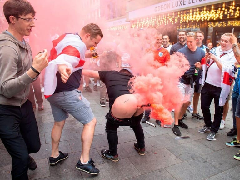 Performer in crouched position releases red smoke flare as audience watches during outdoor Fringe show.
