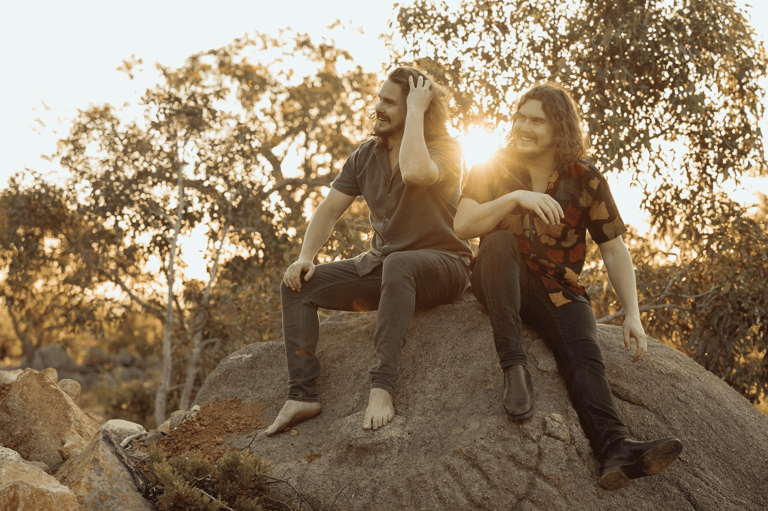 Two people sit on a large rock outdoors at golden hour, backlit by trees.