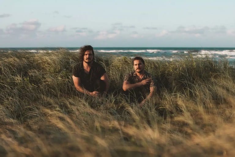 Two people seated in coastal grass dunes with ocean and sky visible behind them.