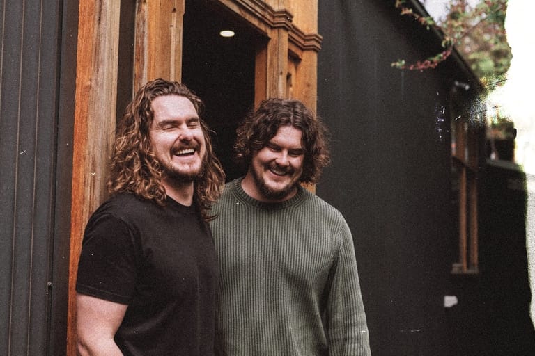 Two men with curly hair stand together outside a wooden-framed doorway, smiling at the camera.