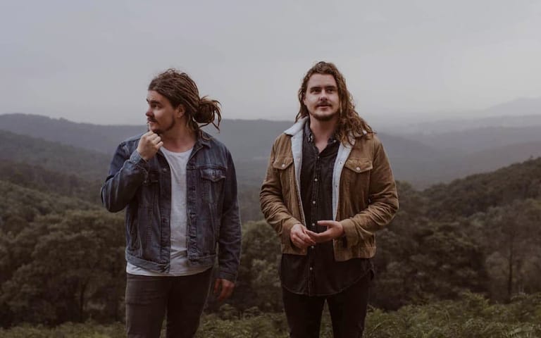 Two men with long hair stand in a misty mountain landscape wearing casual jackets.