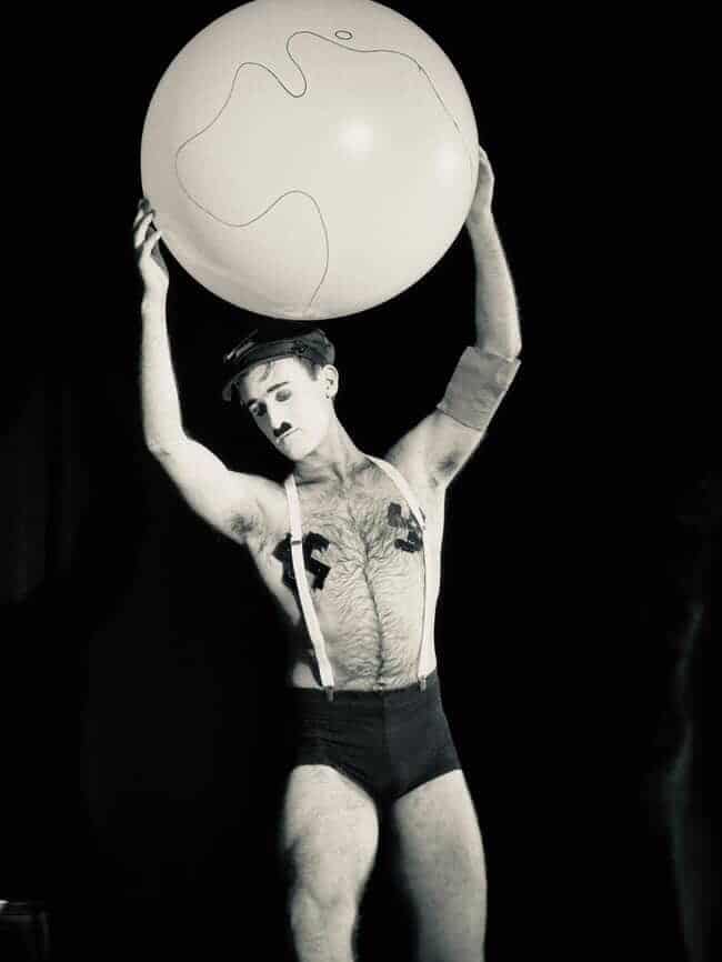 Performer holds a large white sphere overhead in a theatrical pose against a dark background.