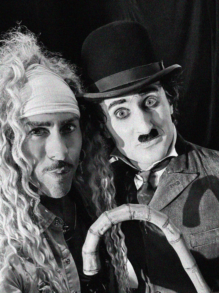 Two performers in character makeup and period costume pose together against a dark background in a theatrical production.