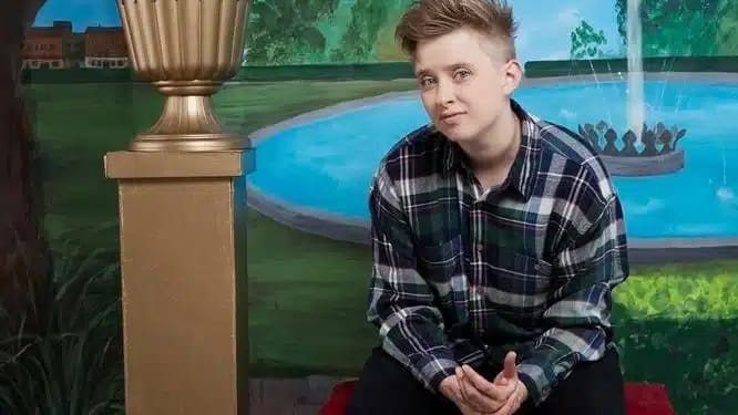 Young person in navy plaid shirt sits beside a gold trophy during an interview setting.
