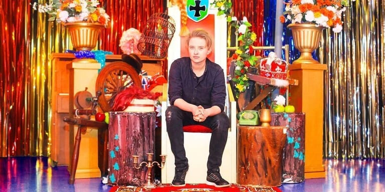 Person seated on stage surrounded by colourful decorative elements and gold curtaining during an interview.
