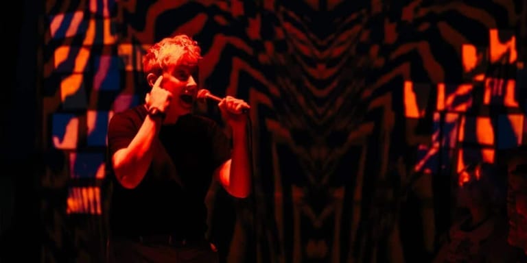 Performer singing into microphone under warm orange stage lighting with geometric backdrop.