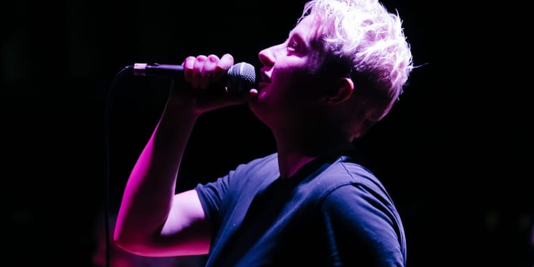 Person with white hair singing into a microphone against a dark background lit by purple and blue stage lights.