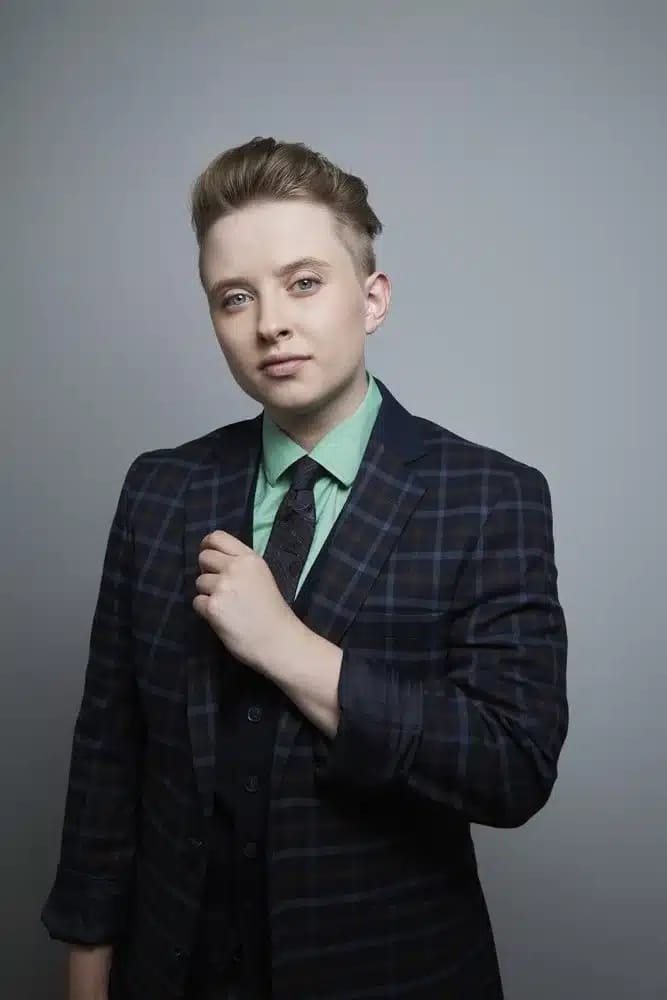 Person in navy plaid blazer and green shirt against neutral grey background, hand near shoulder in portrait pose.