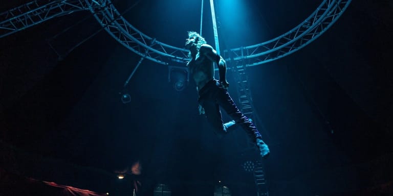 Aerial performer suspended mid-air beneath industrial rigging against a dark, moody stage backdrop.
