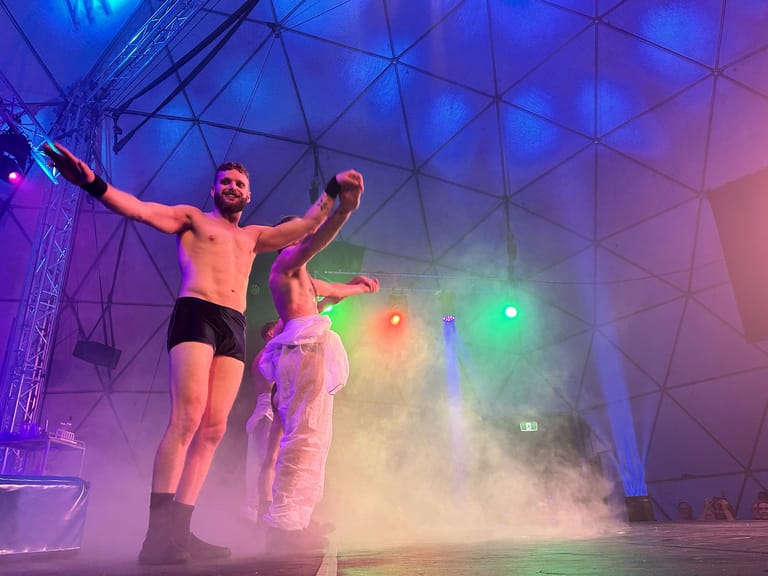 Shirtless performer with arms raised stands amid coloured stage lights and fog, with a second performer visible beside him.