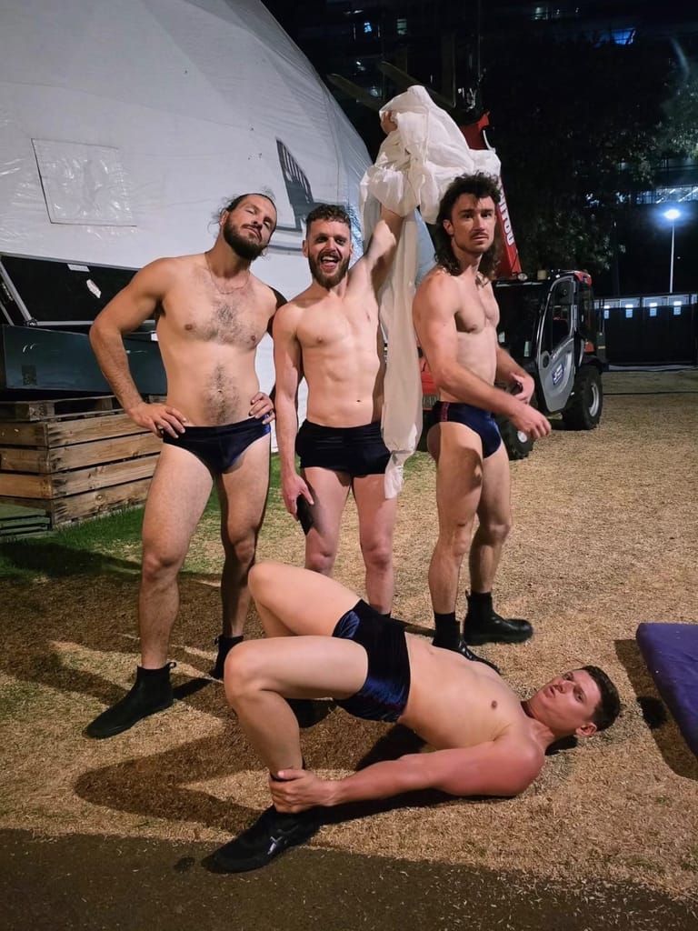 Four shirtless male performers and one performer lying on the ground pose outside a caravan during an outdoor theatre production.