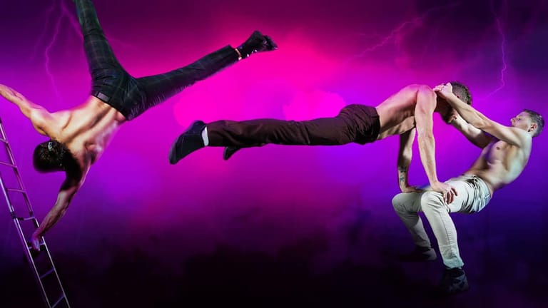 Two aerialists perform an inverted horizontal pose against vibrant magenta stage lighting during a circus performance.