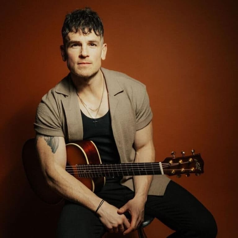 Musician holding an acoustic guitar against a warm orange backdrop, wearing a casual shirt and black undershirt.