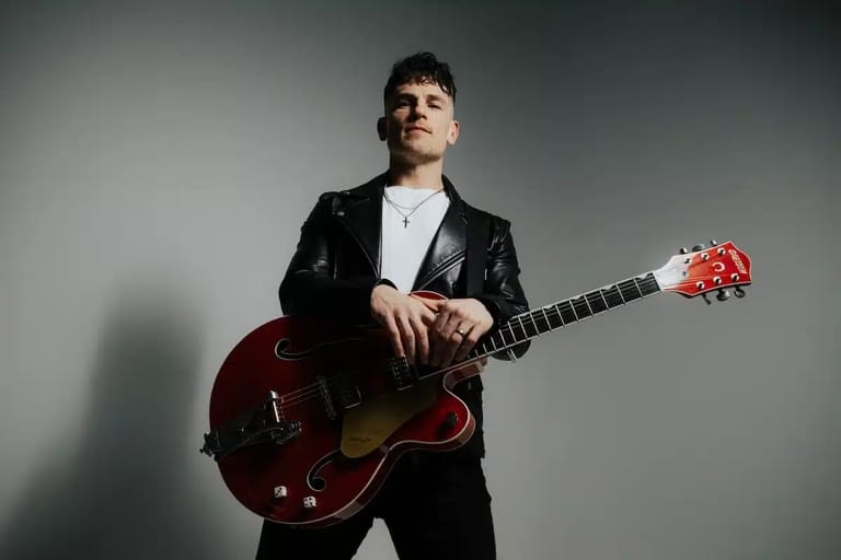 Musician in black jacket holding a dark red semi-acoustic guitar against a neutral grey background.