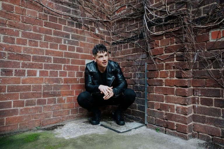 Man in black jacket crouching against red brick wall in intimate venue setting.