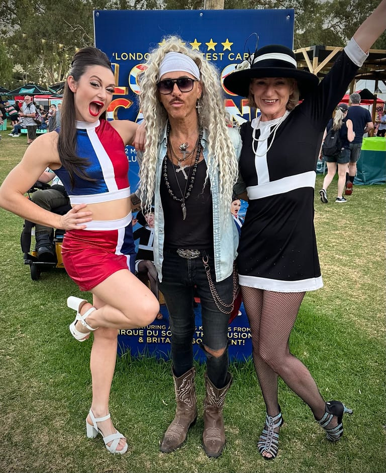Three performers in theatrical costumes pose together on a grassy outdoor stage area during the Adelaide Fringe festival.