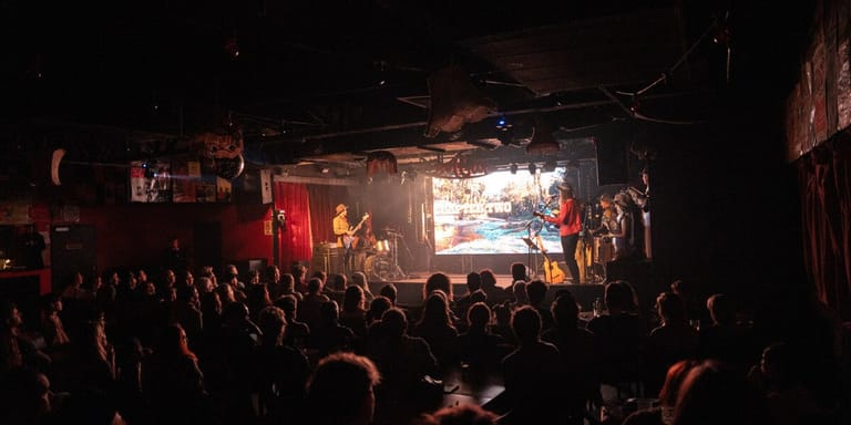 Silhouetted crowd facing a brightly lit stage with performers during a live music event.