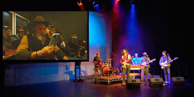 A live band performs on stage with coloured lighting while a close-up of a vocalist appears on a projection screen above.