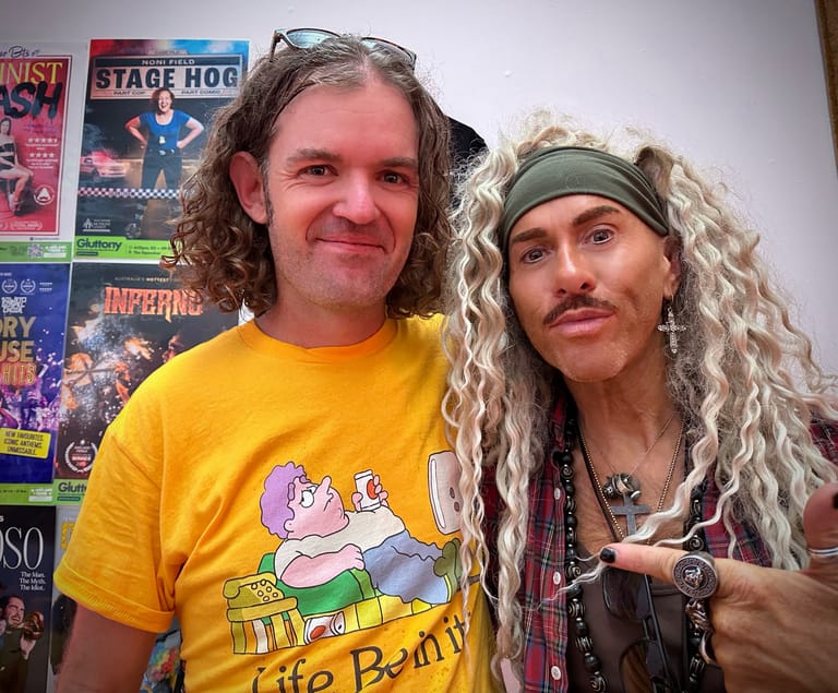 Two performers in colourful costumes and wigs pose together against a backdrop of concert posters and memorabilia.