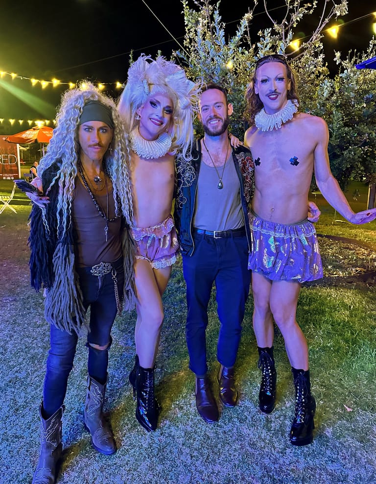 Four performers in elaborate cabaret costumes with wigs and ruffled collars pose outdoors under festive string lights.