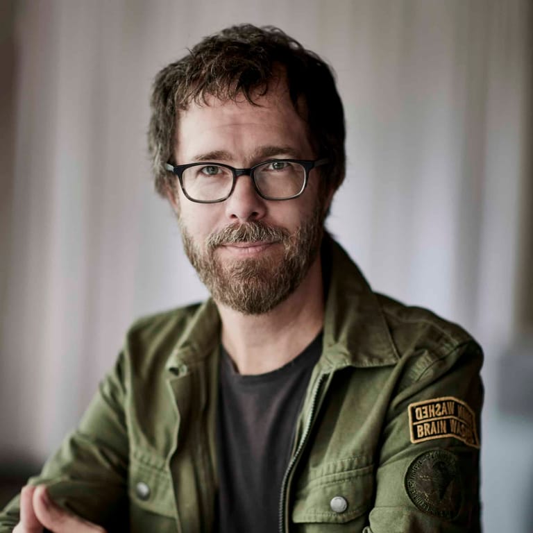 Man with glasses and beard wearing olive-green jacket against neutral background.