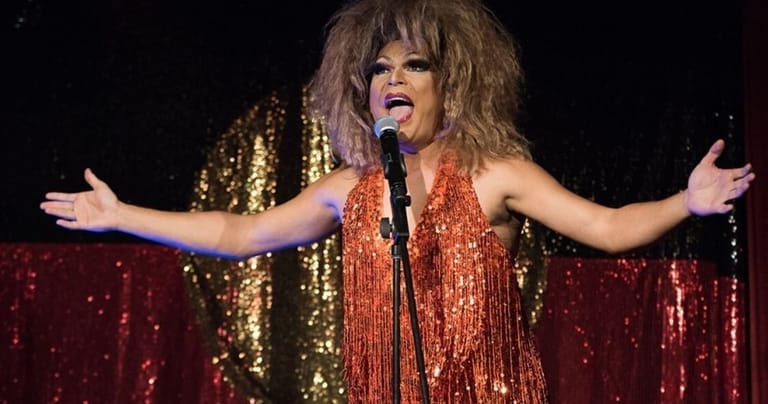 Performer in a shimmering red-and-gold sequined dress with arms outstretched on a sparkling stage.