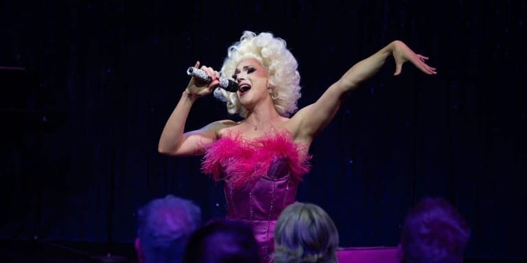 Performer in a hot pink feathered dress sings into a microphone with one arm extended, lit by purple stage lighting.