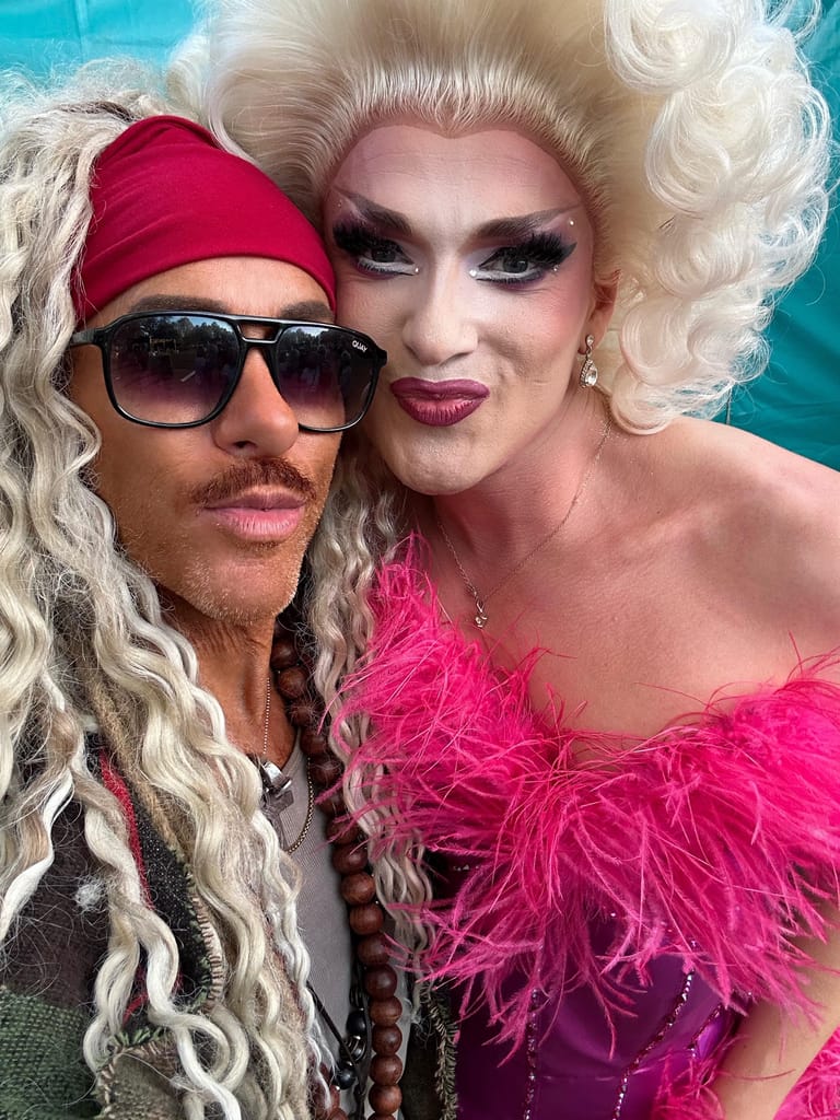 Two performers in elaborate drag costumes with voluminous white wigs and bold makeup pose together against a turquoise background.