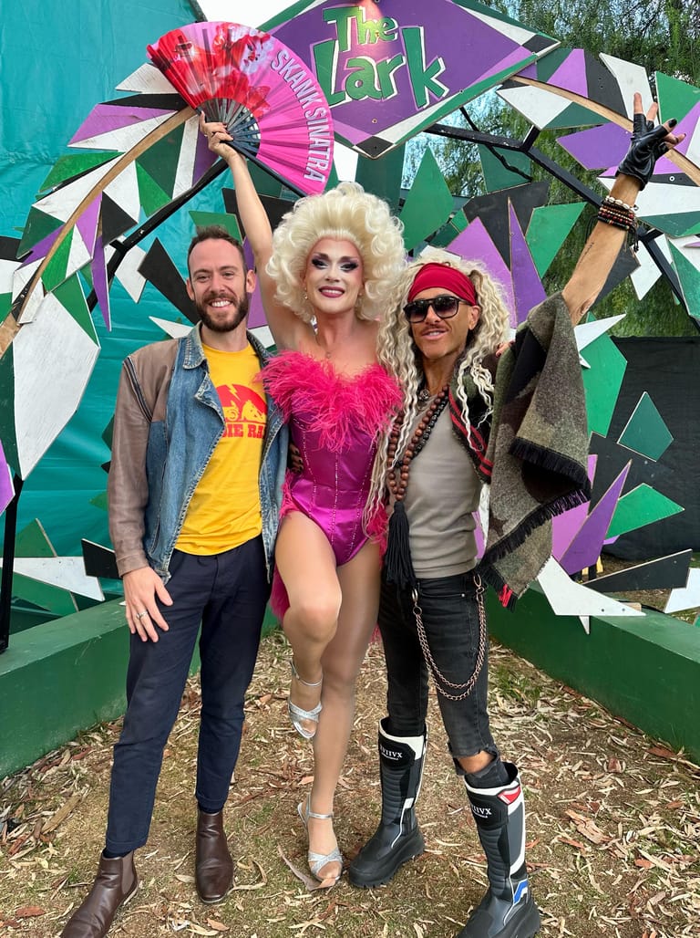 Three performers in vibrant drag costumes pose before a colourful geometric mural backdrop.