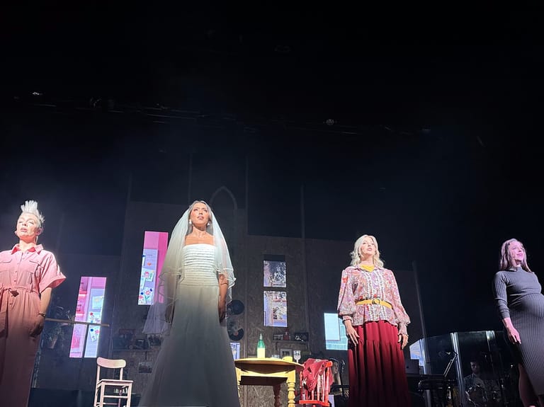 Four performers stand on a darkly lit stage set with neon-lit geometric structures and furnishings during a theatrical production.