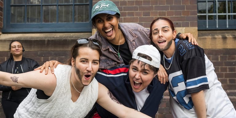 Four young performers pose together against a brick wall, smiling and gesturing enthusiastically at the camera.
