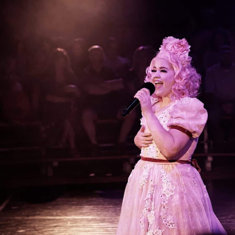 Performer in elaborate pink costume and voluminous wig sings into microphone on stage, audience visible in background.