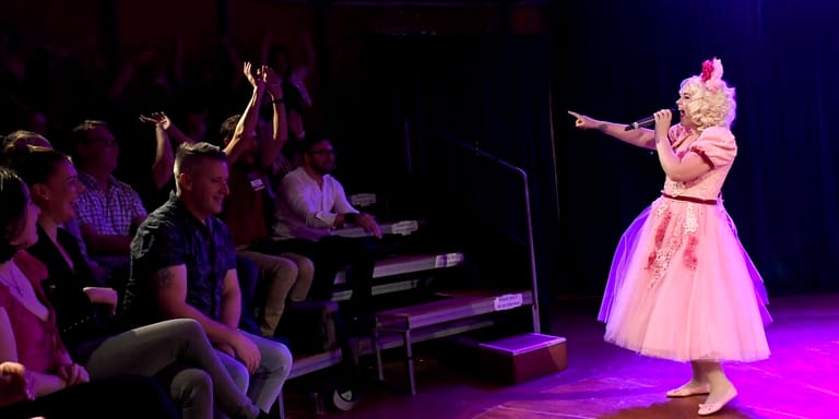 Performer in pink dress interacts with seated audience members under purple stage lighting during a theatrical production.