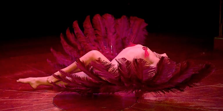 Dancer in flowing magenta costume reclines on stage floor amid dramatic red lighting.