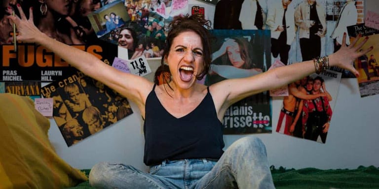 Performer with arms outstretched laughing in front of a wall covered with 90s memorabilia and posters.