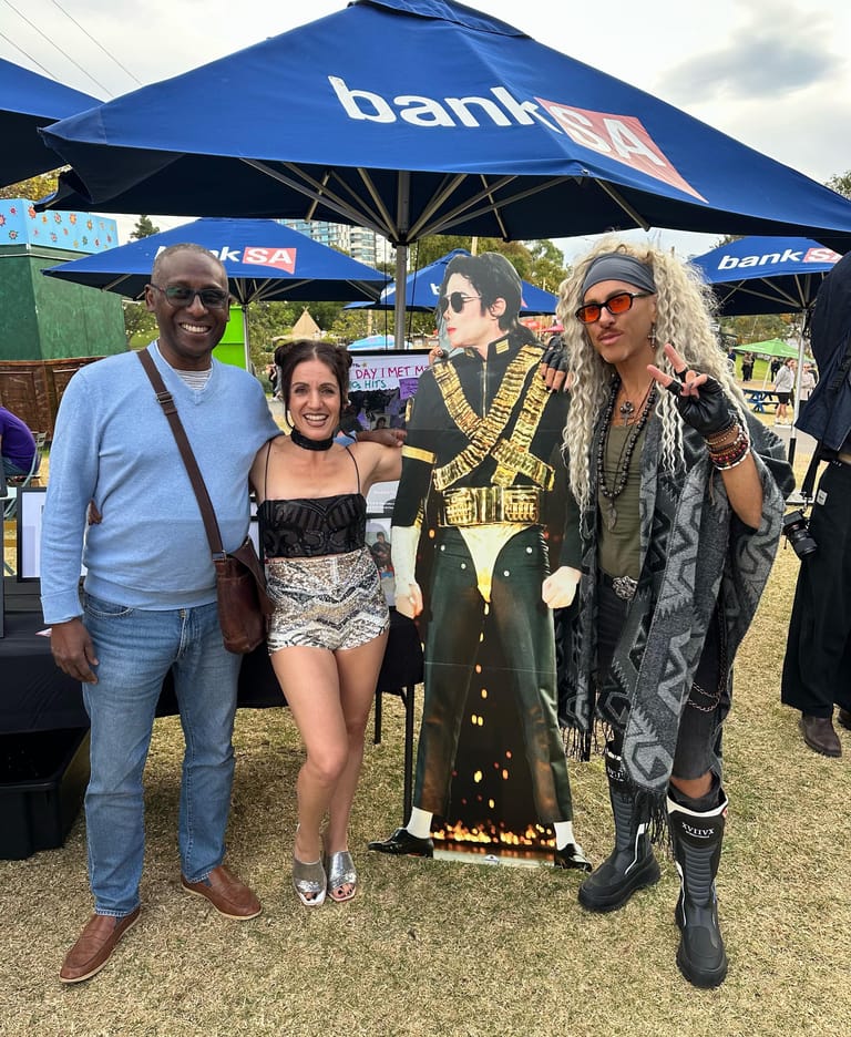 Two attendees pose with a Michael Jackson tribute figure dressed in his iconic gold-trimmed black jacket under a blue bank umbrella.