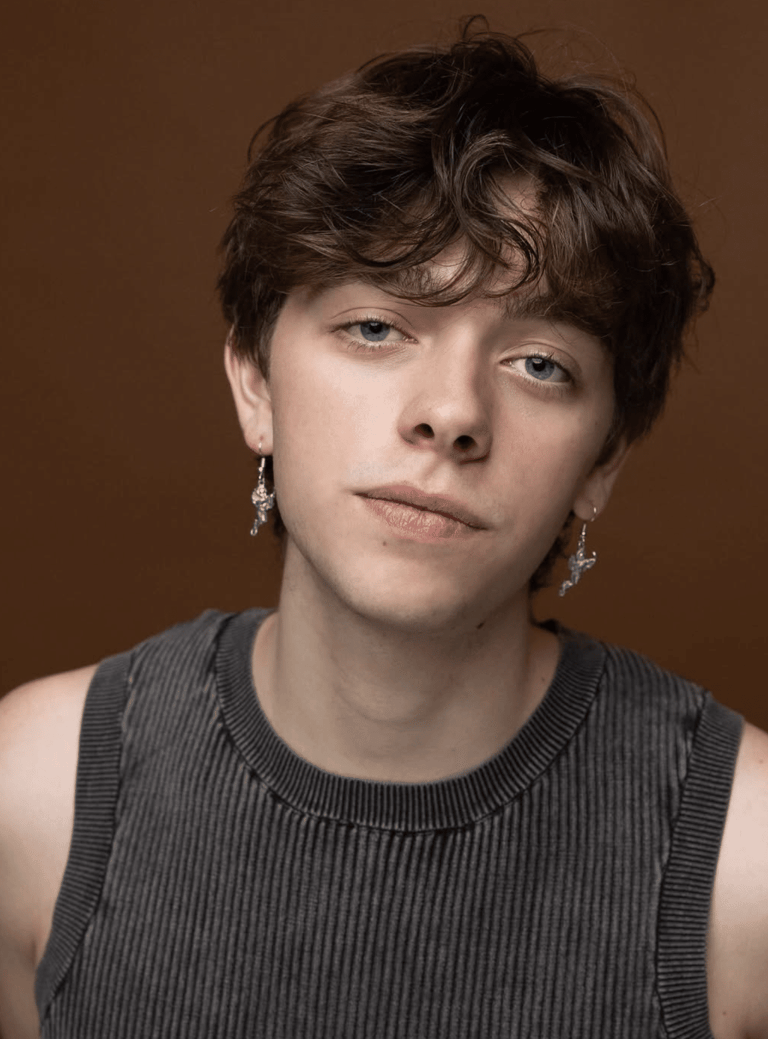 Person with short dark hair and dangling earrings wearing a grey ribbed sleeveless top against a brown background.