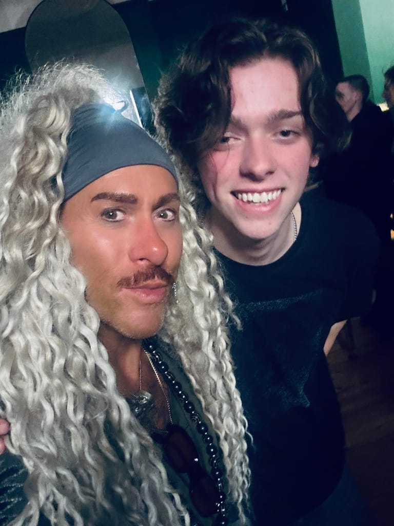 Two performers pose together at a comedy show, one wearing a long blonde curly wig and studded outfit, the other in dark clothing.