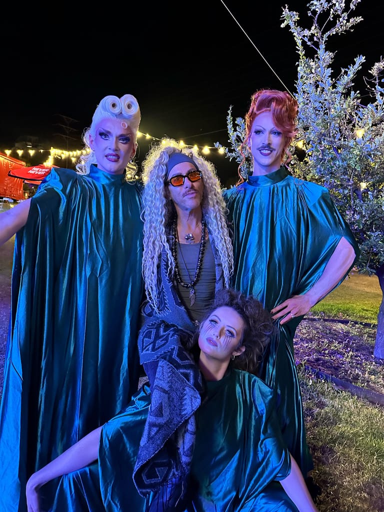 Four performers in blue-lit costumes pose against illuminated trees at an outdoor venue at night.