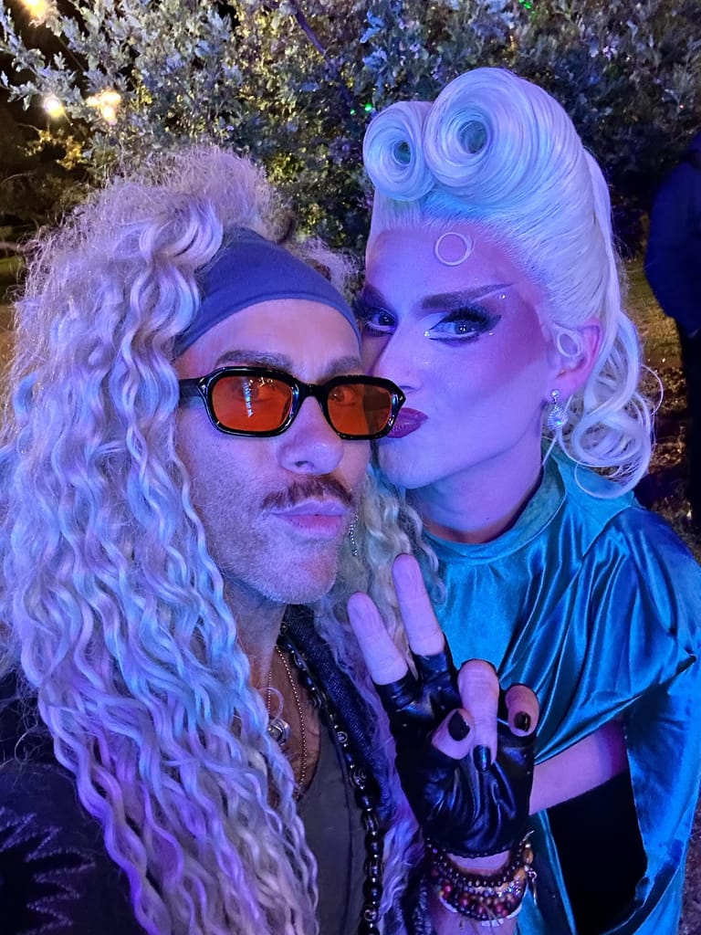 Two performers in blonde wigs and bold makeup pose against a glittering backdrop under purple stage lighting.
