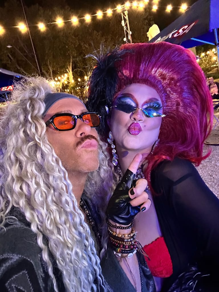 Two performers in dramatic makeup and wigs pose at an outdoor Adelaide Fringe venue lit by string lights.